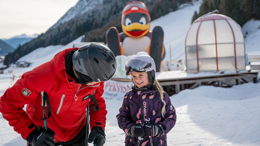 Ein Skilehrer erkundigt sich nach dem Wohlbefinden der kleinen Kursteilnehmerin bei den Planberg- & Wiesenliften in Pertisau. Im Hintergrund Pinguin BOBO.